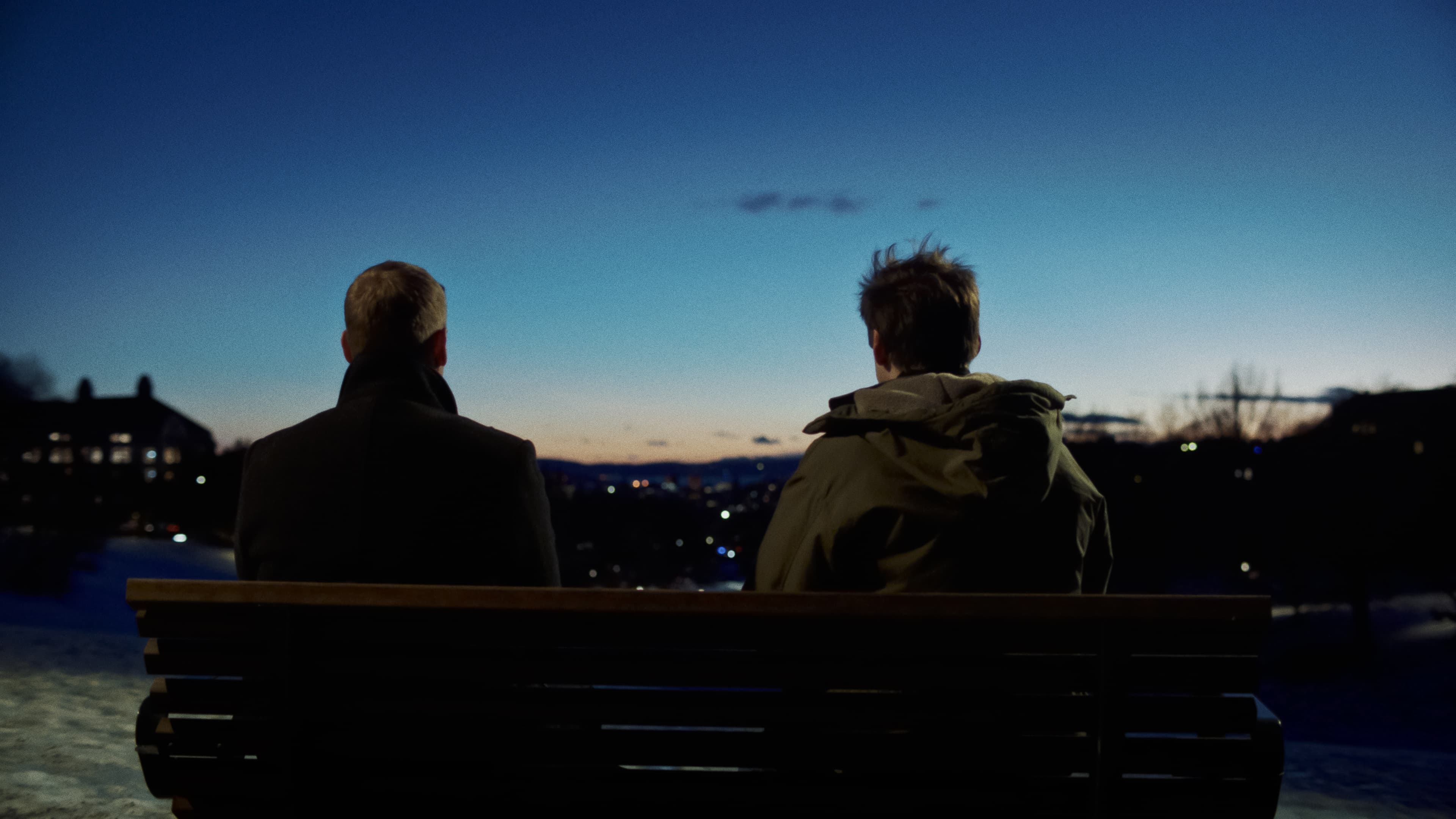 To personer sitter på en benk i skumringen, vendt bort fra kameraet og med blikket vendt mot et bylandskap med fjerne lys under en dypblå himmel.