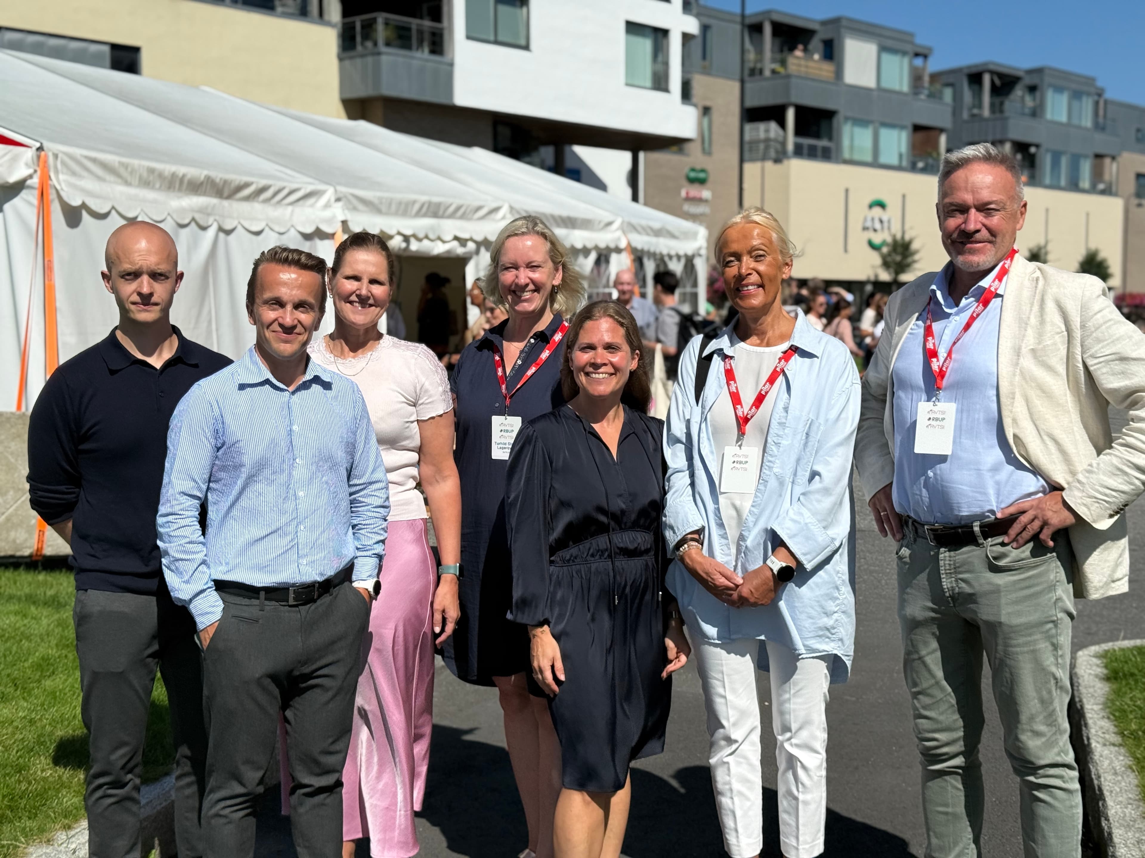 Sju voksne mennesker, fire kvinner og tre menn, står ute en solrik dag og smiler til kameraet. De er kledd i business-antrekk og har på seg event-badges. Et hvitt telt og moderne bygninger skimtes i bakgrunnen.