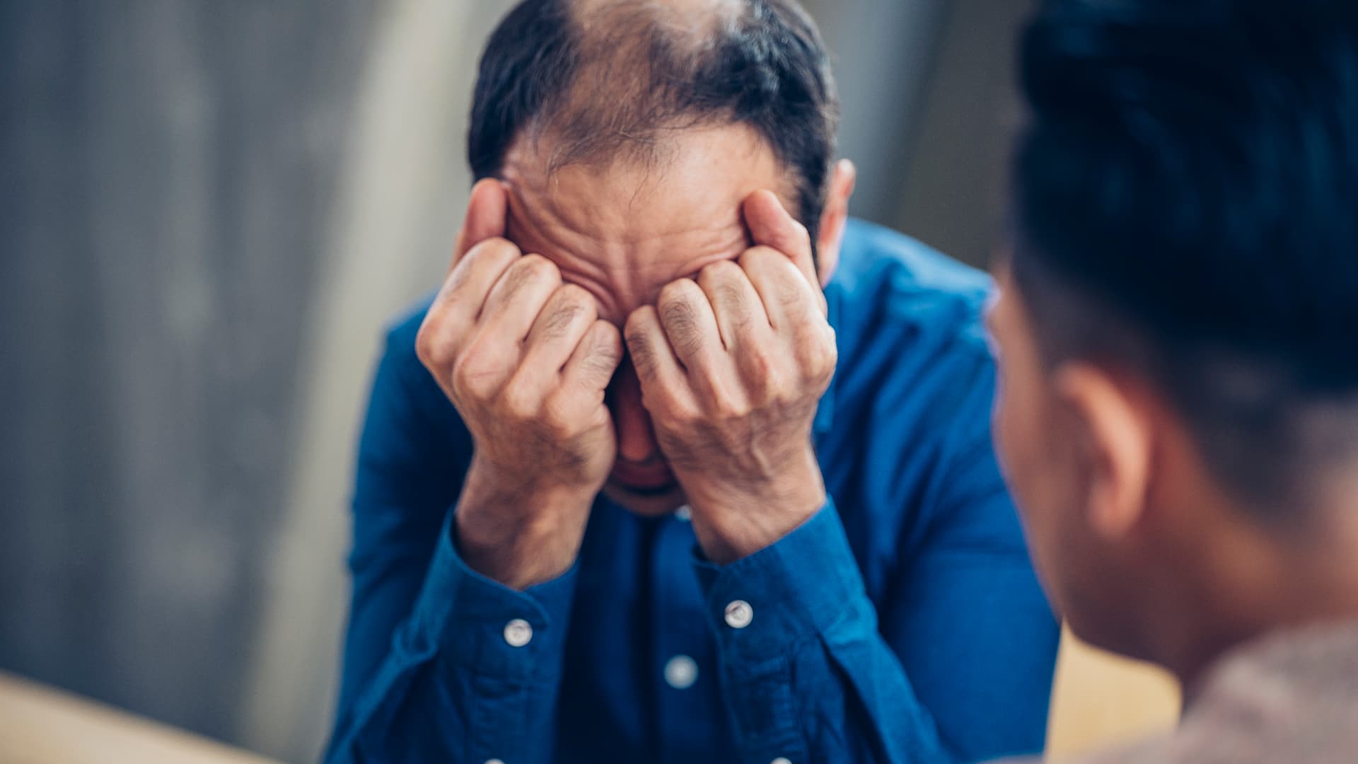 En mann sitter med hodet i hendene, synlig fortvilet, mens en annen person sitter i nærheten vendt mot ham, noe som antyder et øyeblikk med emosjonell støtte eller samtale.