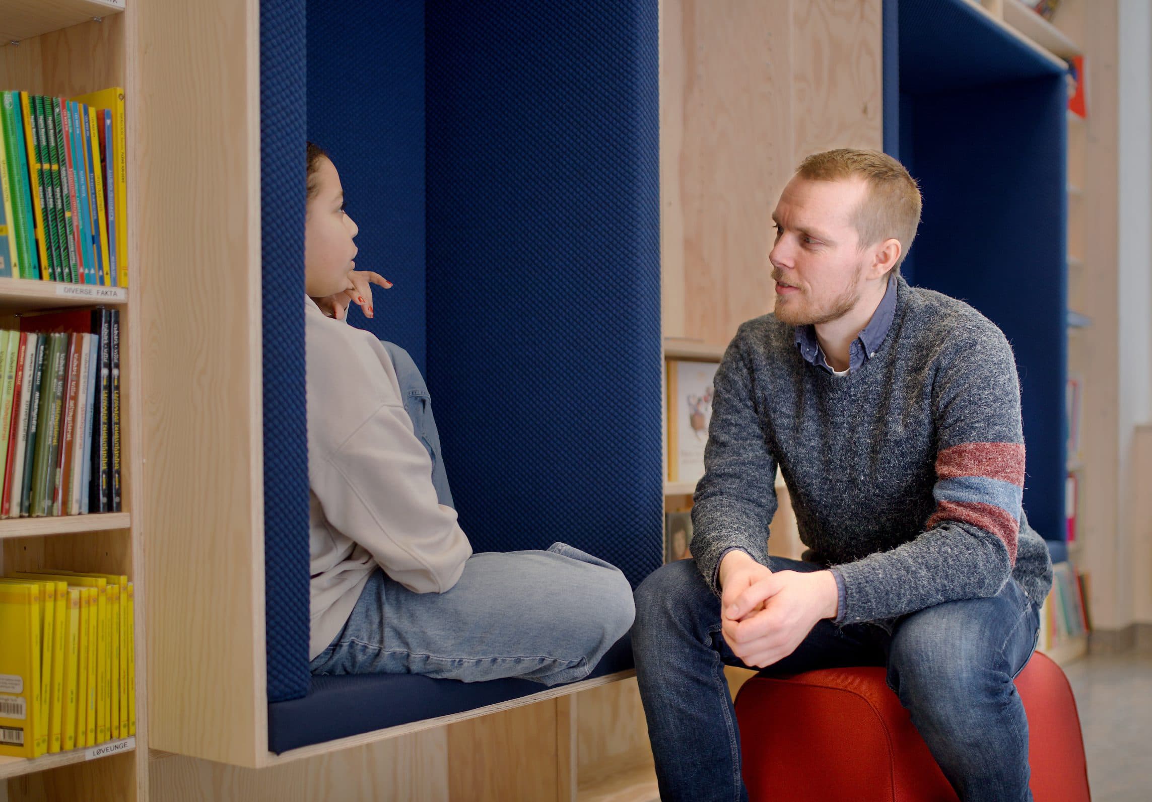 Et barn sitter i en polstret alkove, vendt mot en voksen som sitter på en rød krakk. Hyller med bøker er synlige til venstre.