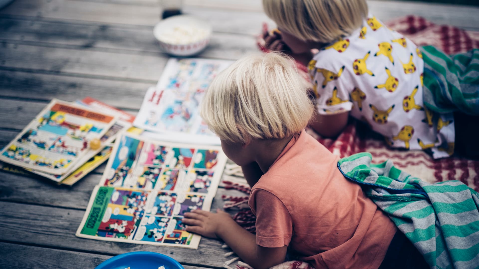 To små barn med blondt hår ligger på et teppe utendørs og leser fargerike tegneserier sammen. En skål med snacks og en blå tallerken står ved siden av på treflaten.