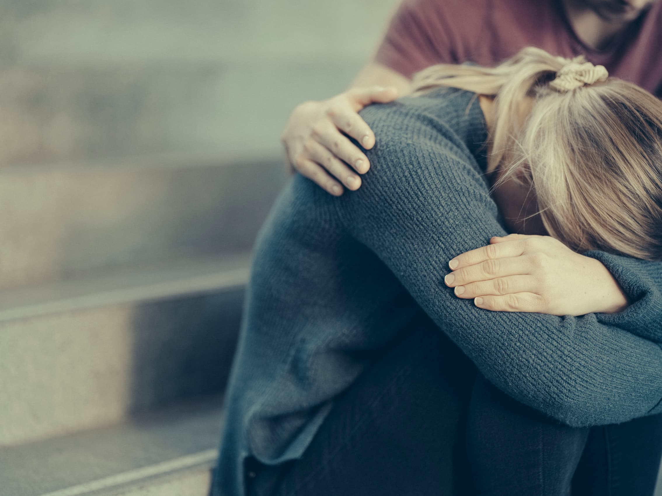 En person sitter på trapper, hodet ned, med en annen person som trøster dem ved å legge en hånd på skulderen.